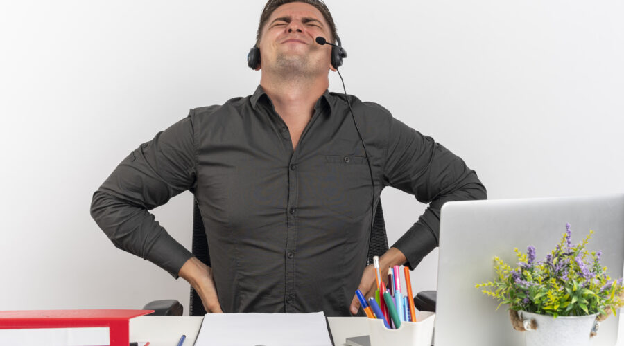 young annoyed blonde office worker man on headphones sits at desk with office tools using laptop holds his back looking up isolated on white background with copy space Tutto d’un tratto avverto dolore. Ma perché? Genesi riabilitazione san giovanni lupatoto